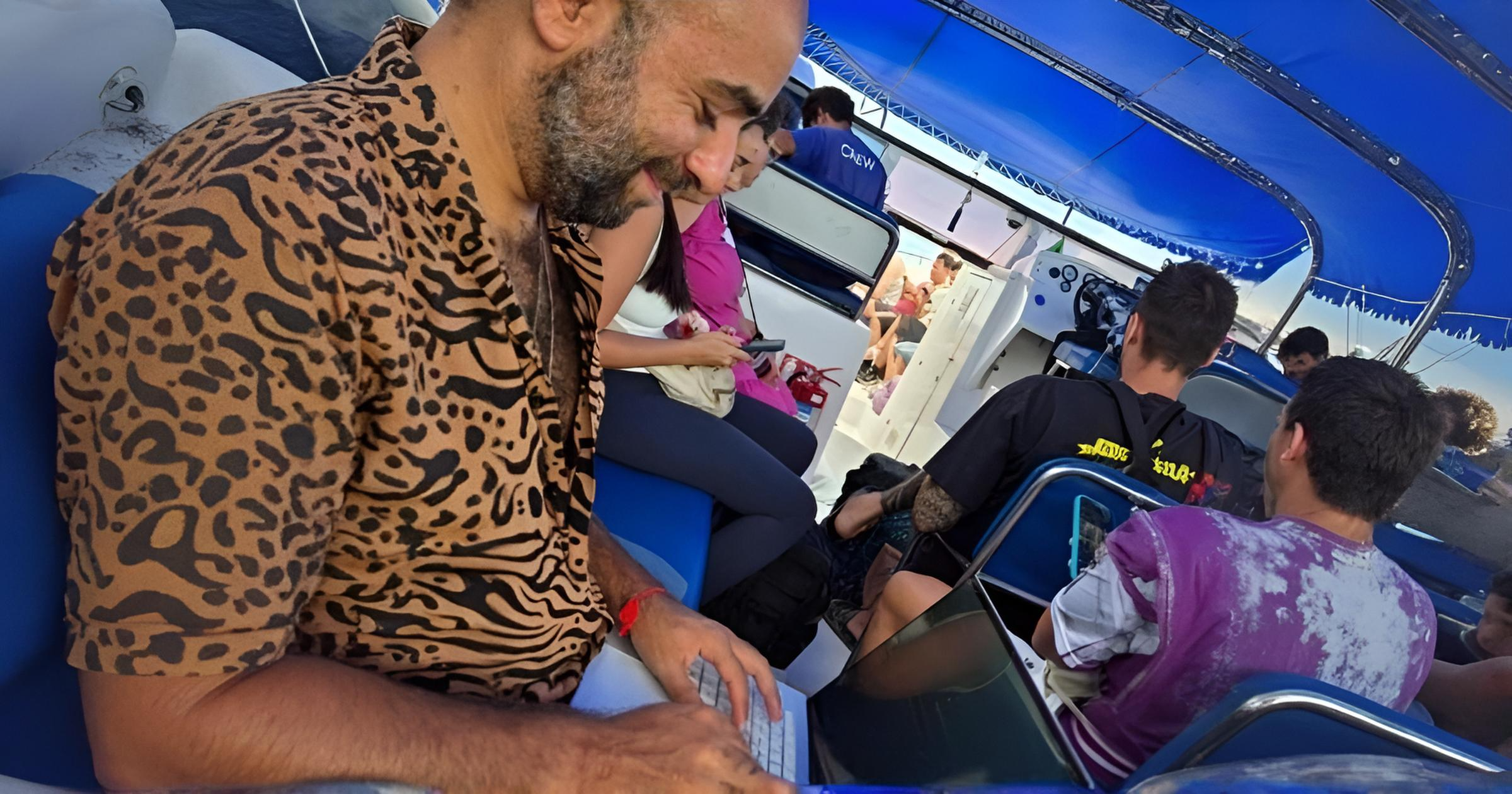 אדם יושב וכותב במחשב נייד על סירה לצד נוסעים נוספים.